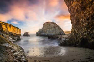 This scene, depicted near Santa Cruz, California, has a lot of calming, tranquil drama to it. It seems like a place where a Pirate might try to spend the night, before heading back out to sea the next day. The dramatic orange sunset, and large rock formation of Shark Fin Cove, make this Fine Art Photograph a great composition of land, sea and sky. Clark Schiring took this image.